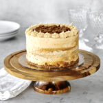 Lotus Biscoff cake on a brown wooden cake stand with glasses, plates, and silverware also in the scene
