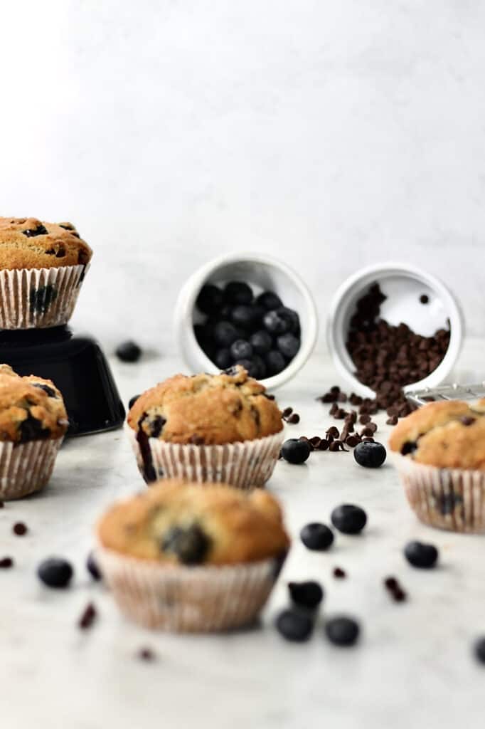 blueberry and chocolate chip muffins scattered on a countertop, surrounded by fresh blueberries and mini chocolate chips