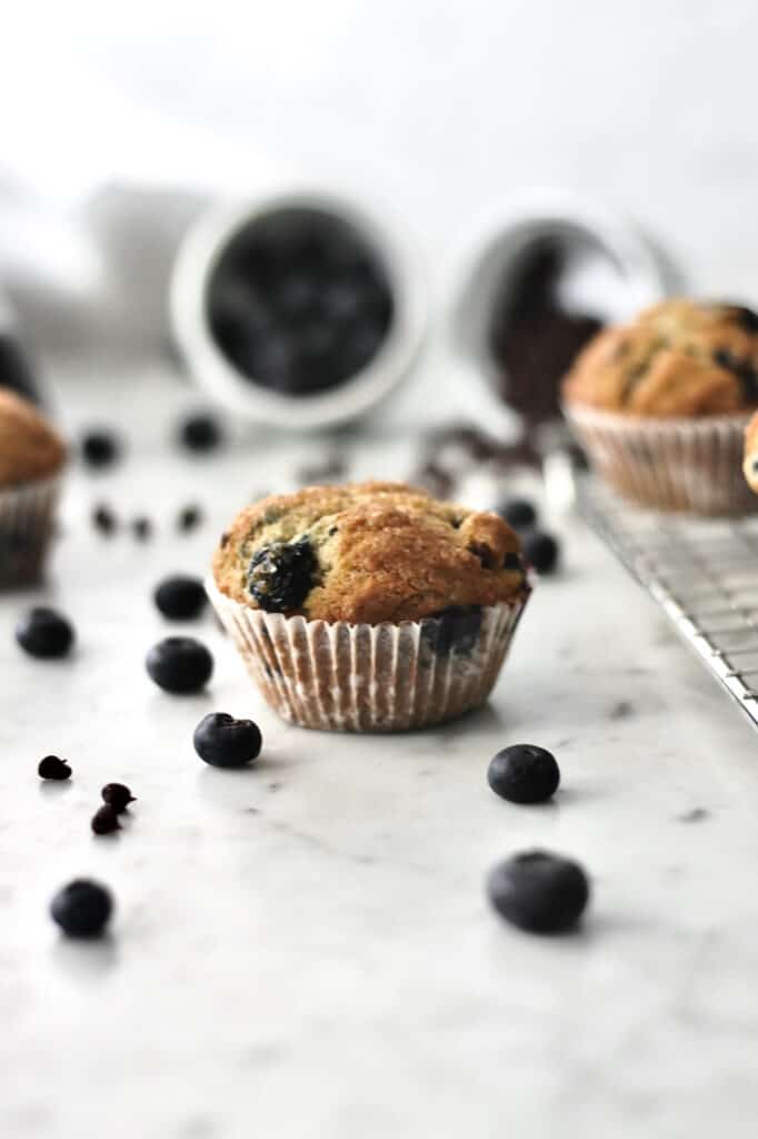 muffin on a countertop, surrounded by mini chocolate chips and fresh blueberries