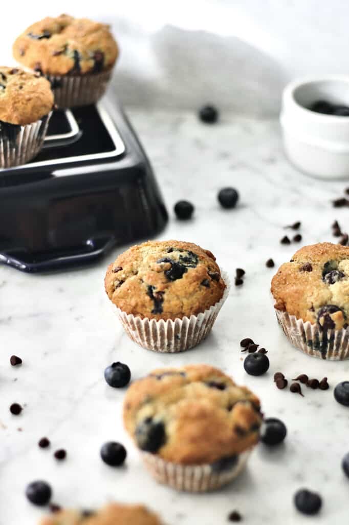 muffins on a countertop, surrounded by mini chocolate chips and fresh blueberries