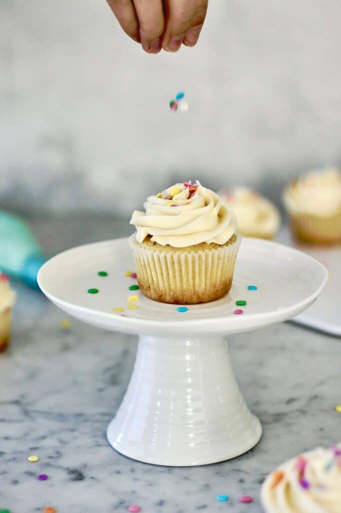 action shot of adding sprinkles to a vanilla cupcake on a white stand, surrounded by lots of confetti sprinkles