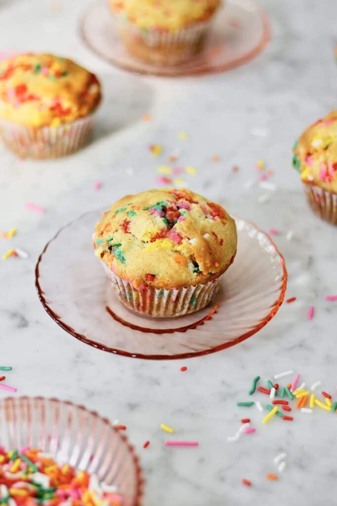 image of a baked funfetti muffin in a white liner sitting on a light pink plate, surrounded by a bowl of sprinkles, sprinkles on the counter, and a few muffins scattered throughout the image in the background