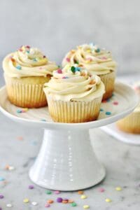 three vanilla cupcakes on a small white cake stand, surrounded by sprinkles