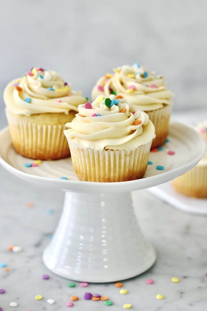 three vanilla cupcakes on a small white cake stand, surrounded by sprinkles