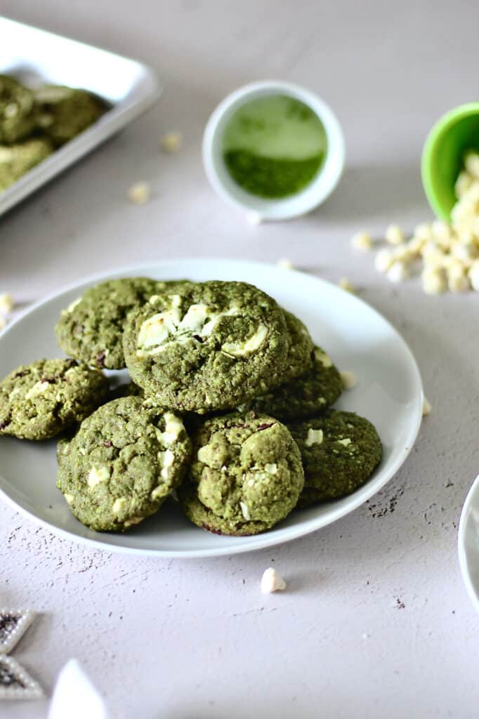 bright green colored cookies full of white chocolate chips, sitting on a white plate with matcha powder and more white chocolate chips in the background