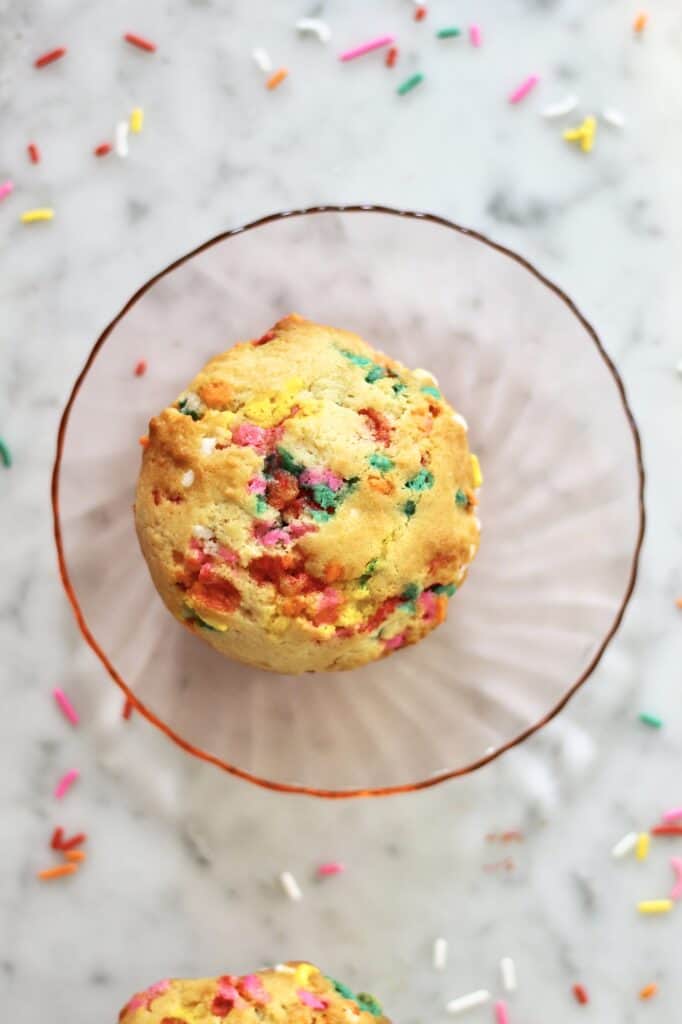 birthday cake muffin sitting on a light pink plate, surrounded by lots of colorful sprinkles