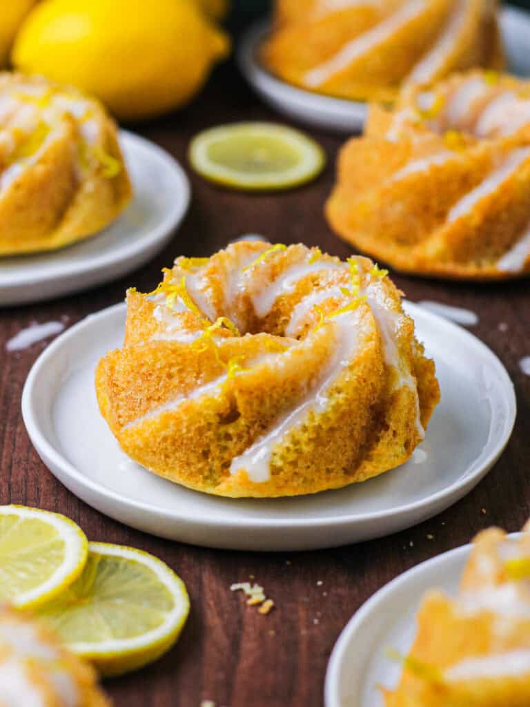 A wooden countertop with mini lemon Bundt cakes sitting in white plates on the surface. They're coated with a light glaze and lemon zest.