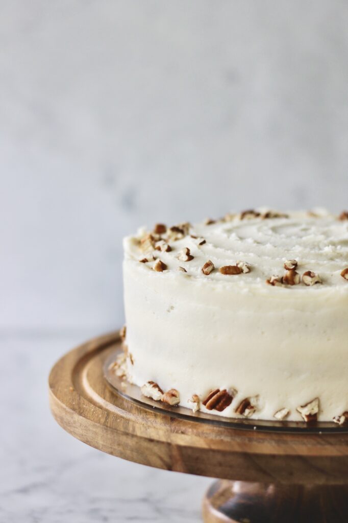 Image of cake for my old fashioned carrot cake recipe with pineapple. Cake is sitting on a brown wooden cake stand, decorated with crushed pecans