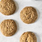 image of a few peanut butter cookies on a grey background with a spoon of peanut butter in one corner and a ramekin full of peanut butter in the diagonally opposite corner