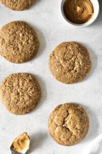 image of a few peanut butter cookies on a grey background with a spoon of peanut butter in one corner and a ramekin full of peanut butter in the diagonally opposite corner