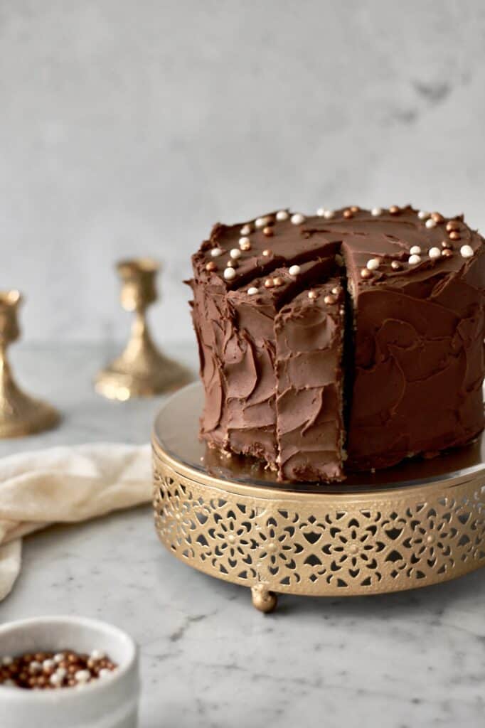 image of banana cake with chocolate frosting, sitting on a short gold cake stand, with gold and white sprinkles decorating its top and sitting in a ramekin in the corner of the photo
