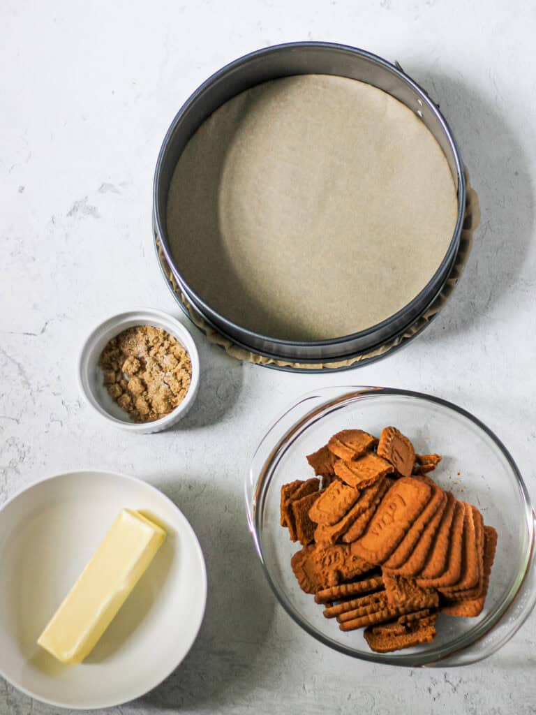 Process shot for Biscoff pie crust recipe, showing Biscoff cookies, stick of butter, and brown sugar in separate bowls, next to a springform pan on a grey background.