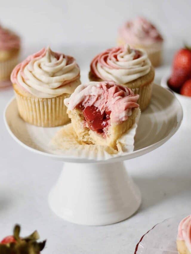 three strawberry filled cupcakes on a white cake stand, one with a bite into it so you can see the inside of the cupcake