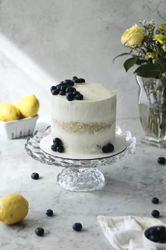 white lemon and blueberry cake on glass cake stand with blueberries on top of it