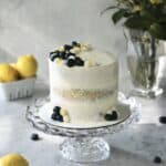 lemon berry cake sitting on glass cake stand, with lemons and blueberries surrounding it