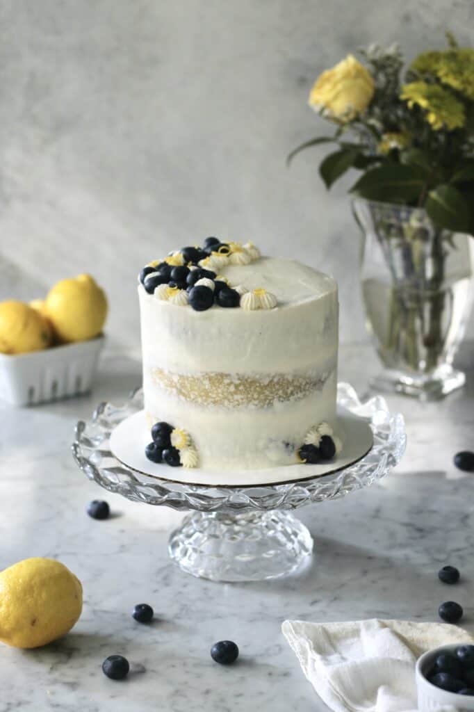 lemon berry cake sitting on glass cake stand, with lemons and blueberries surrounding it