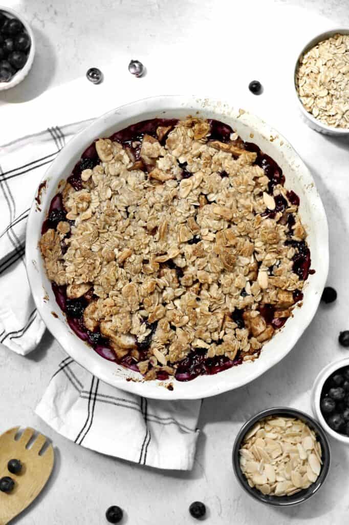 blueberry pear crisp, surrounded by berries and ramekins filled with ingredients for this dessert