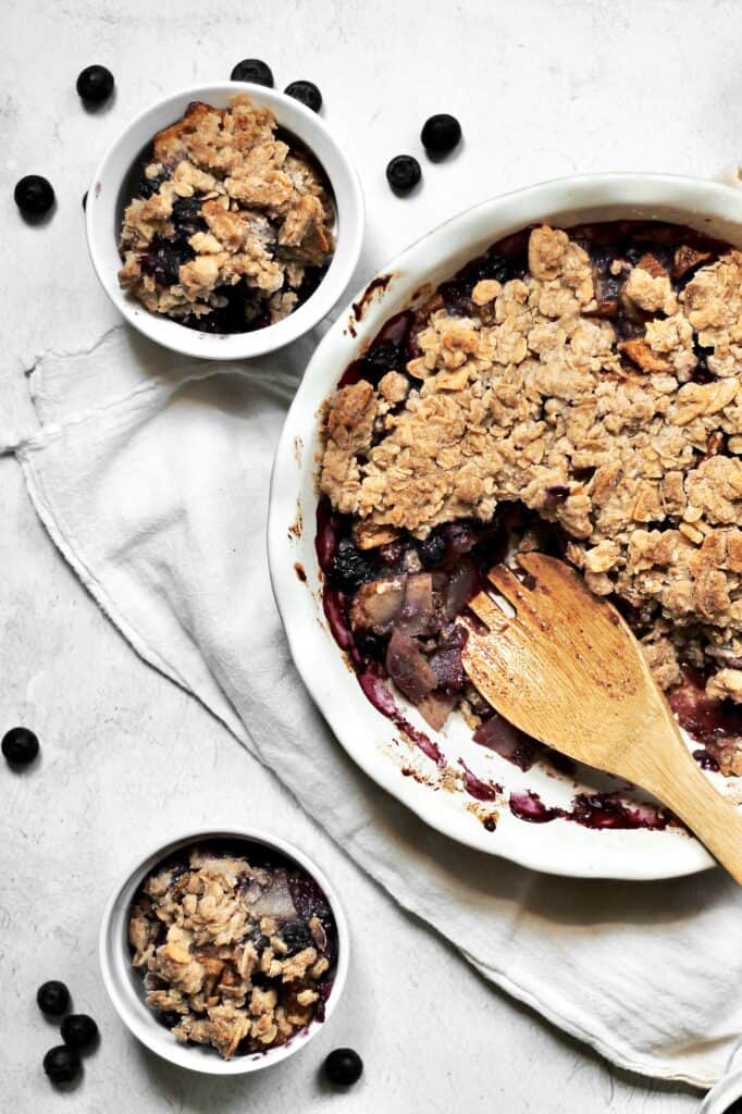 overhead shot of blueberry pear crumble sitting on a white cheesecloth on a grey background, with two ramekins full of servings