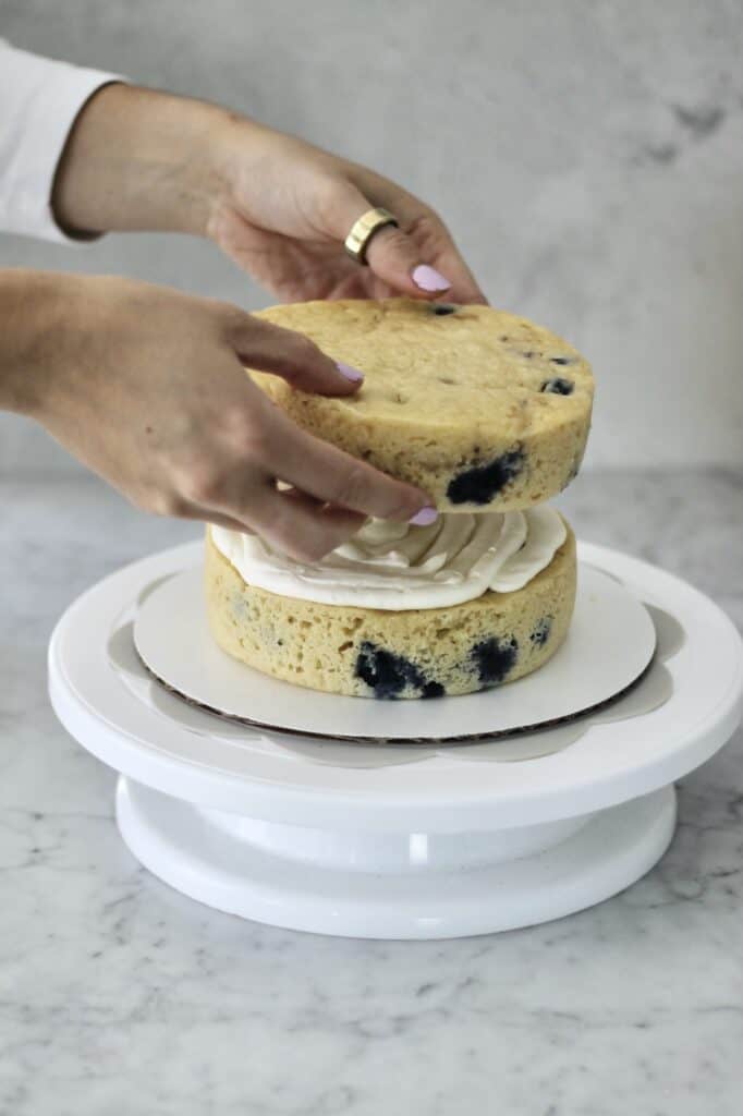 cake turntable with two layers of lemon and blueberry cake on it; image shows baker adding second layer