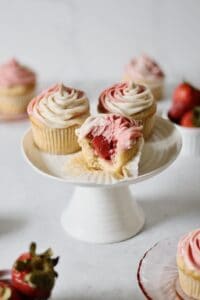 three strawberry filled cupcakes on a white cake stand, one with a bite into it so you can see the inside of the cupcake