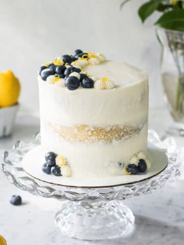 White mascarpone frosting on a lemon layer cake filled with blueberries. The cake is sitting on a glass cake stand, with lemons and berries scattered around it.