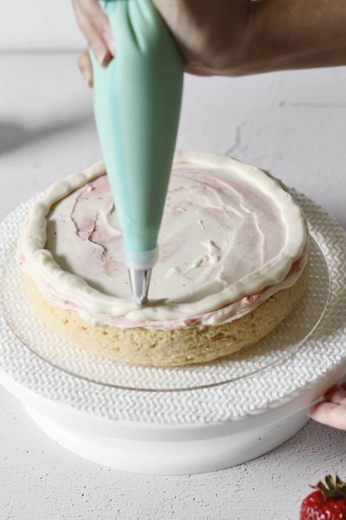 piping frosting onto first layer of vanilla cake with strawberry filling, sitting on a cake turntable