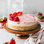 Strawberry mascarpone cheesecake sitting on the base of a springform pan on a cake stand, surrounded by strawberries.