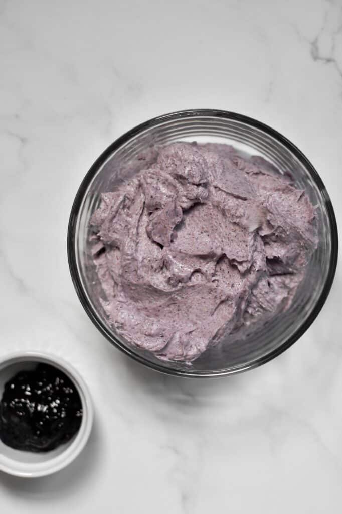 blueberry cake frosting in a clear bowl, next to a few tablespoons of blueberry jam