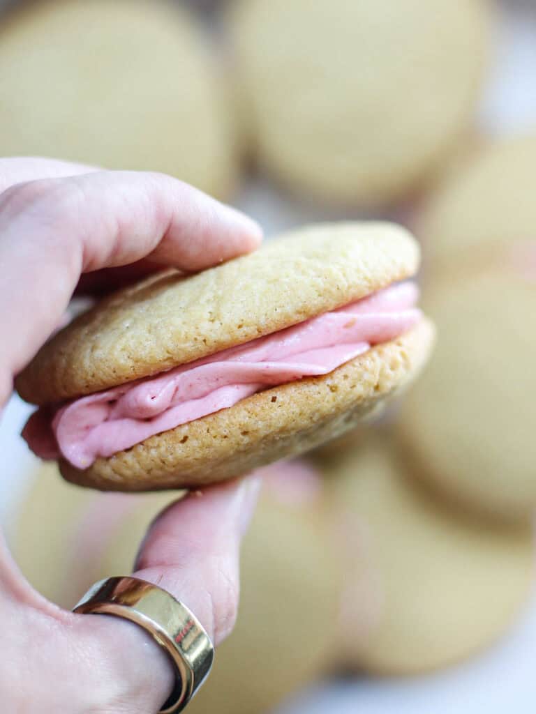 Hand holding a cookie sandwich with pink frosting between the layers.