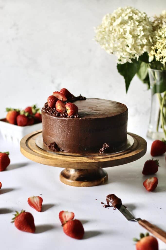 chocolate cake with strawberry filling, sitting on a wooden cake stand, surrounded by strawberries and fresh white hydrangeas in a vase behind it