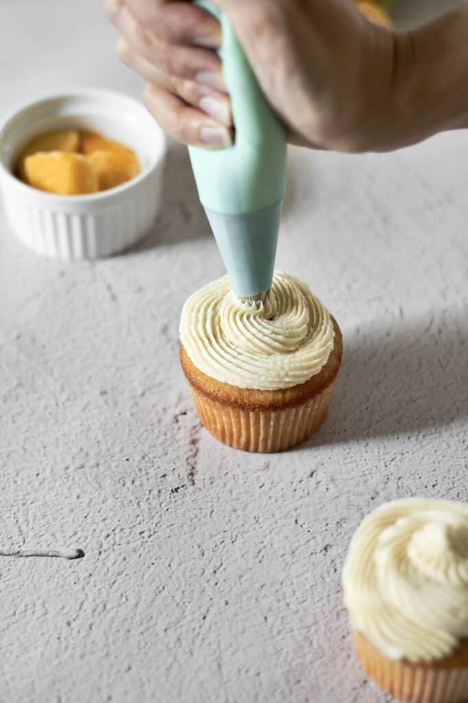 white hand piping mango buttercream onto cupcakes