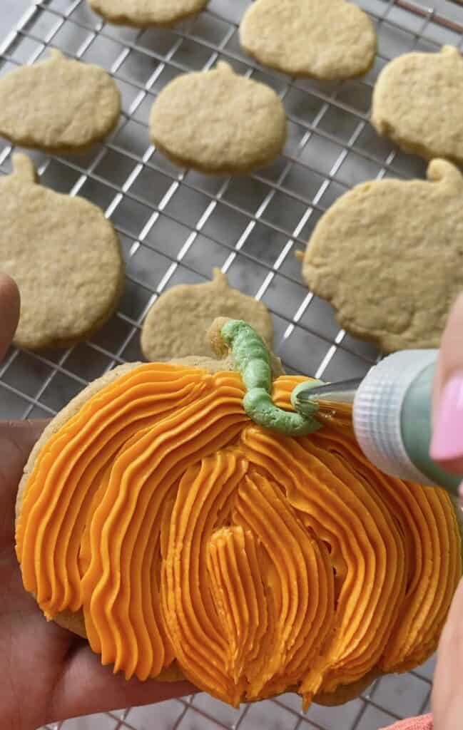 action shot of decorating pumpkin cookies, piping orange frosting onto them
