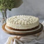 mango mousse cake, sitting in a Biscoff crust on top of a wooden cake stand with a grey background and greenery