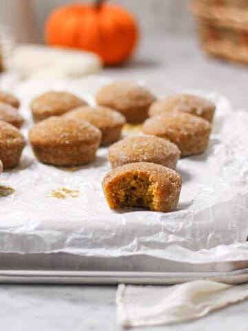 Mini cinnamon sugar pumpkin muffins on parchment paper, one broken open, with pumpkins and a basket behind them.
