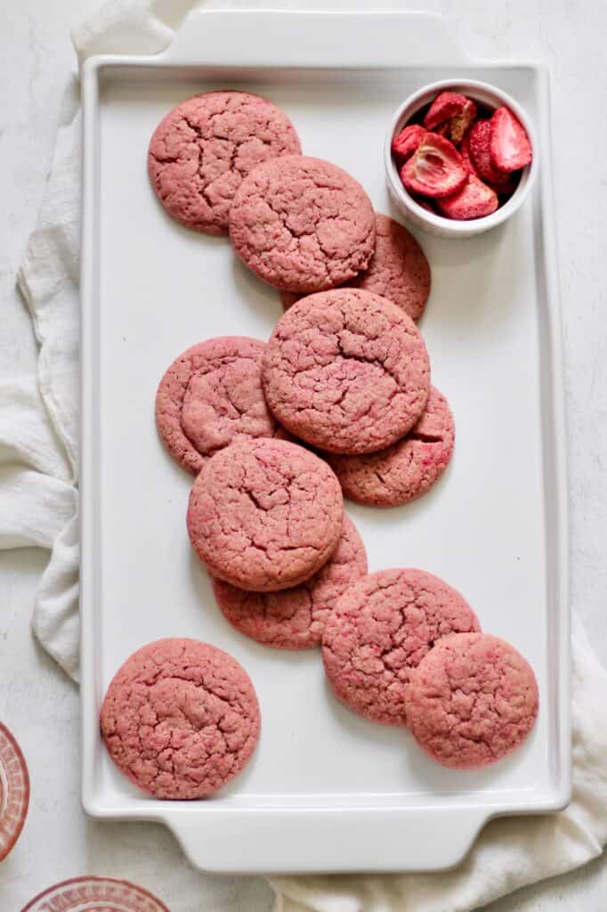 overhead image of pink sugar cookies sitting on a white plate, with freeze dried strawberries in a ramekin in the upper right-hand corner