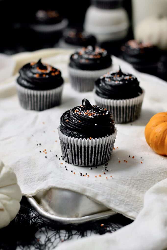 black cocoa cupcakes, sitting on a white linen material in front of a black background and surrounded by sprinkles and mini pumpkins