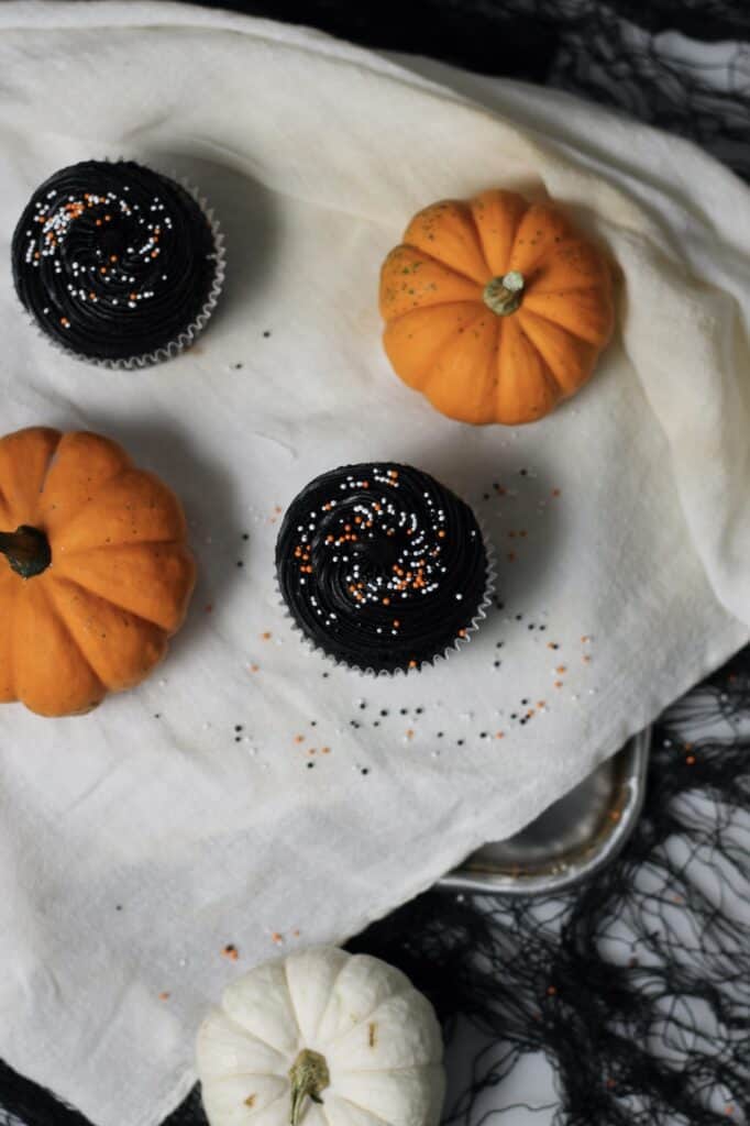 overhead image of black Halloween cupcakes, surrounded by Halloween sprinkles and mini pumpkins