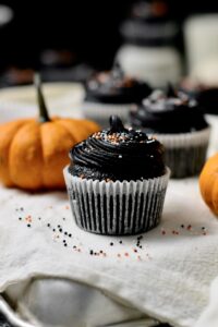 black velvet cupcake in the foreground, sitting on a baking tray, with a pumpkin and more black cupcakes behind it