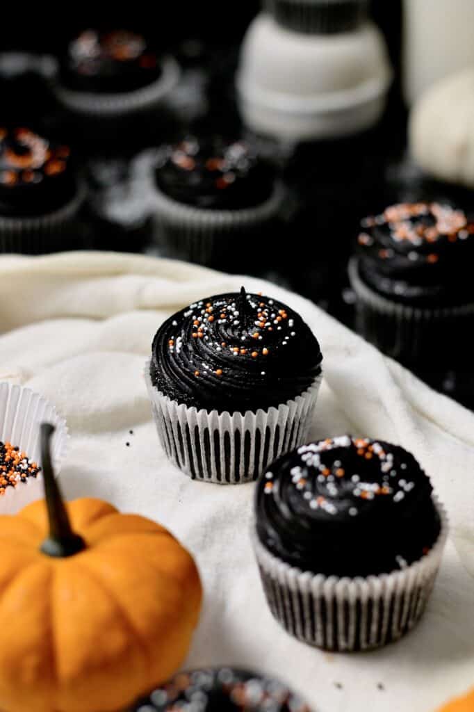 black Halloween cupcakes with orange and white sprinkles, sitting on white linen in front of a mostly black background