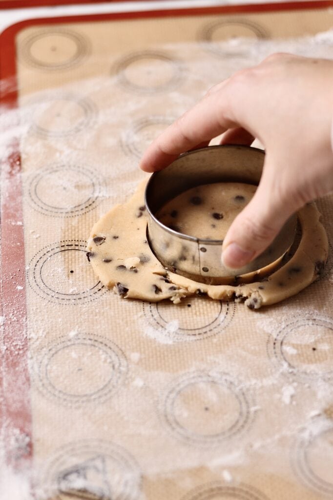 hand using a cookie cutter to cut chocolate chip sugar cookies in round shapes on a silicon mat