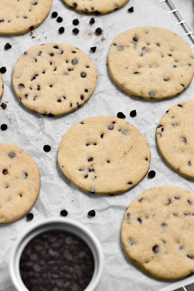 overhead image of unfrosted chocolate chip sugar cookies sitting on a wire rack with parchment paper on top of it and chips scattered throughout