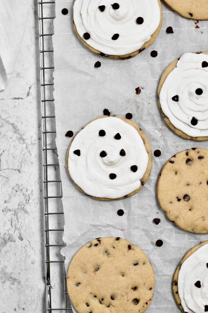 overhead image of frosted and unfrosted chocolate chip sugar cookies sitting on a wire rack with parchment paper on top of it 
