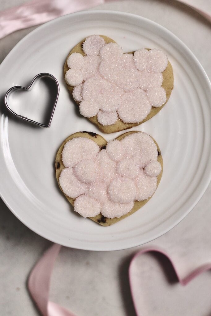 pink frosted cookies with sparkling sugar on top, in the shapes of hearts, sitting on a white cake stand