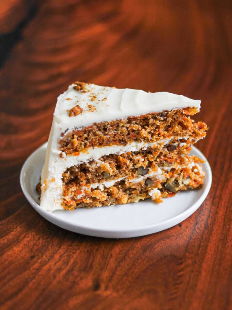 Slice of a carrot cake with cream cheese frosting, sitting on a small white plate against a wooden backdrop.
