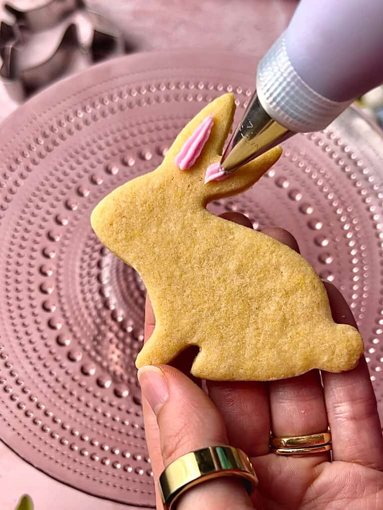 action shot of piping bag decorating a white bunny cookie on a pink background
