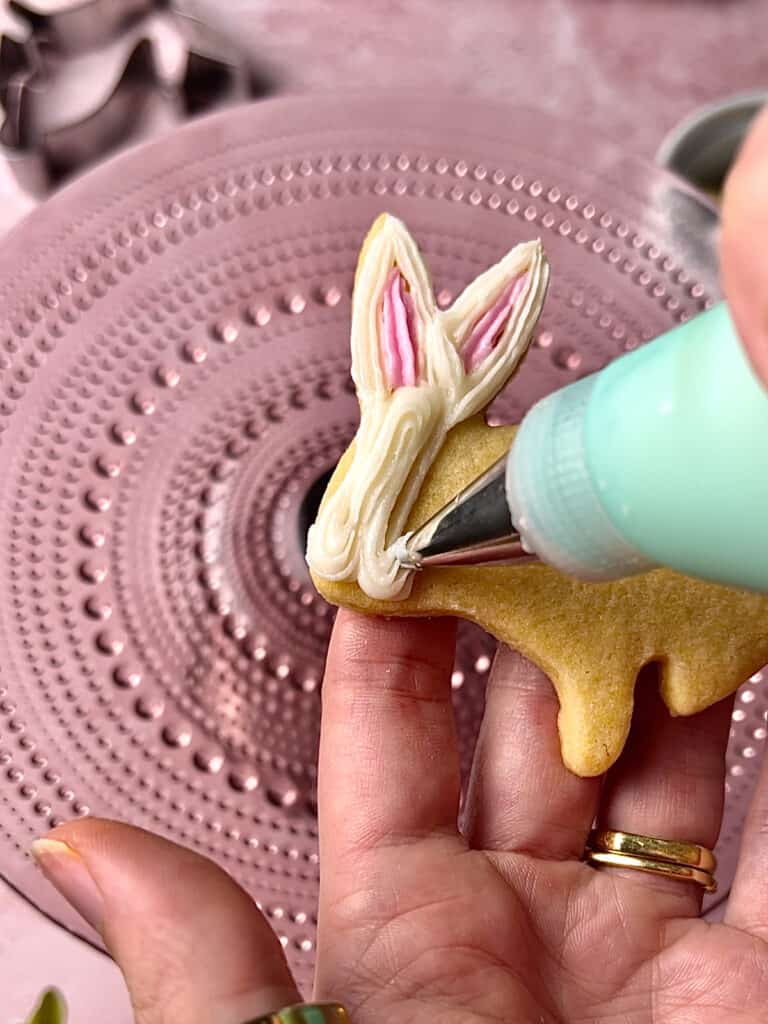action shot of piping bag decorating a white bunny cookie on a pink background