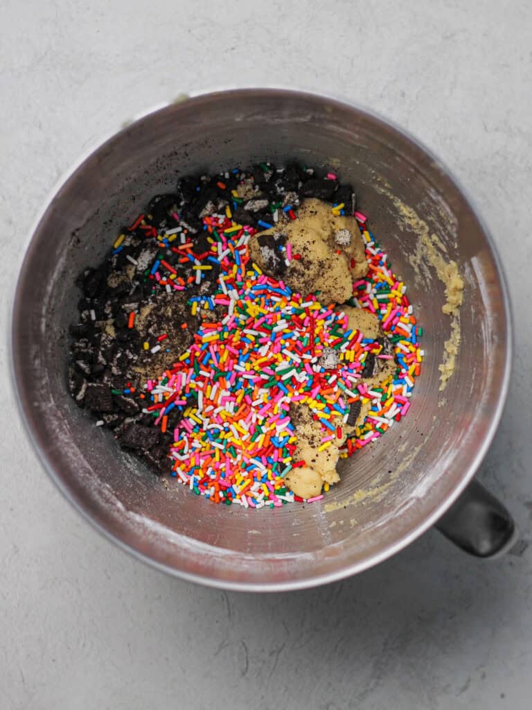 Mixing bowl full of light colored cookie dough, filled with rainbow sprinkles and crushed Oreo pieces.