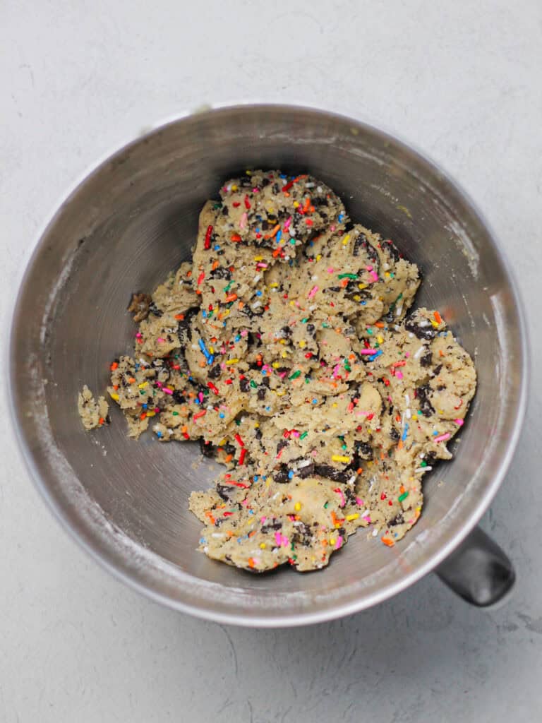 Mixing bowl full of light colored cookie dough, filled with rainbow sprinkles and crushed Oreo pieces.