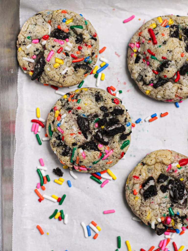Cookies with rainbow sprinkles and Oreo pieces, sitting on a white piece of parchment.