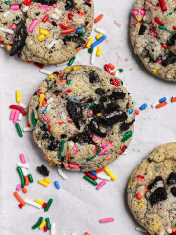 Cookies with rainbow sprinkles and Oreo pieces, sitting on a white piece of parchment.